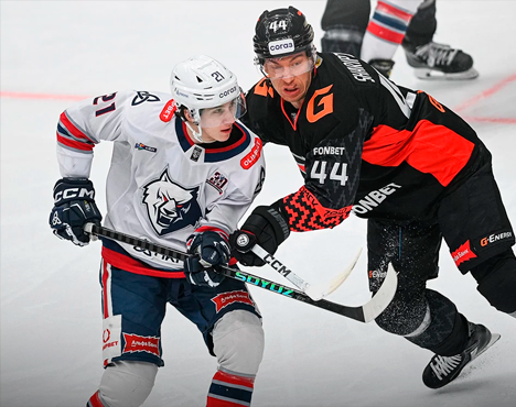 На изображении два хоккеиста в защитной экипировке ведут борьбу за шайбу на льду. Один в белой форме с синими и красными элементами, другой — в черно-оранжевой. Обстановка ледовой арены